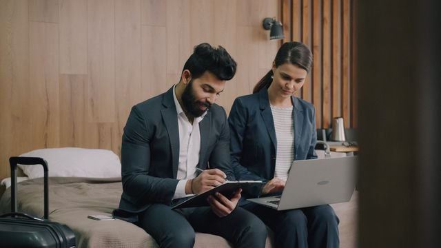 Professionals in formal attire working together in a hotel room, focusing on a laptop and clipboard.